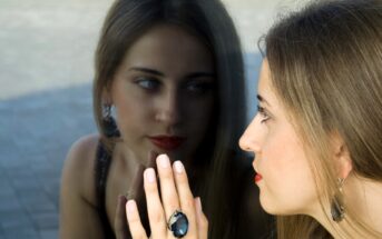 A woman with long hair, wearing bold earrings and a large ring, gazes at her reflection in a glass surface. Her hand is pressed against the glass, and she appears thoughtful, with a slight red lipstick accentuating her features.