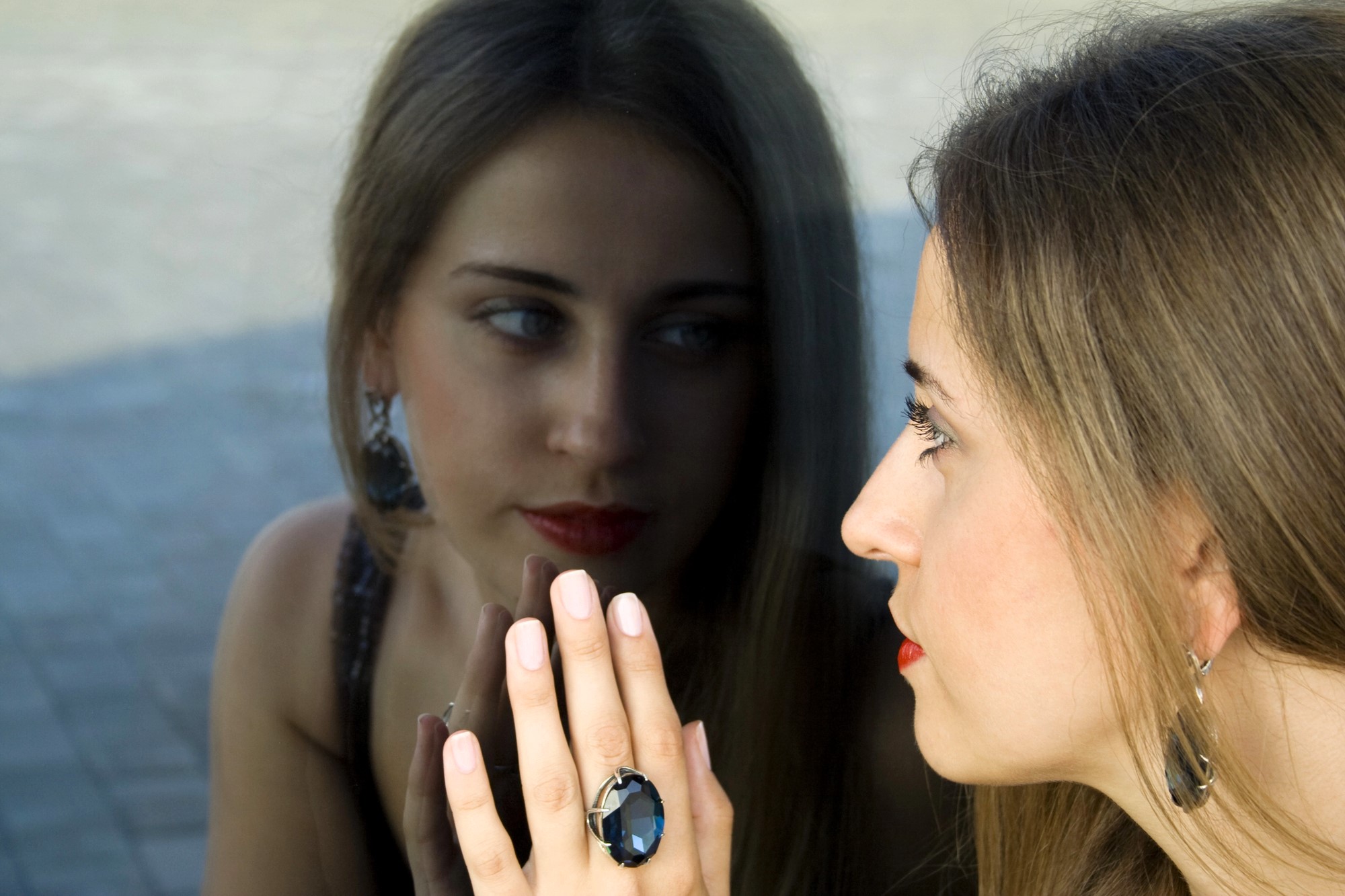 A woman with long hair, wearing bold earrings and a large ring, gazes at her reflection in a glass surface. Her hand is pressed against the glass, and she appears thoughtful, with a slight red lipstick accentuating her features.