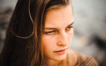 Close-up of a person with long brown hair, looking down and to the side. The background is blurred, highlighting their thoughtful expression and natural skin with light freckles.