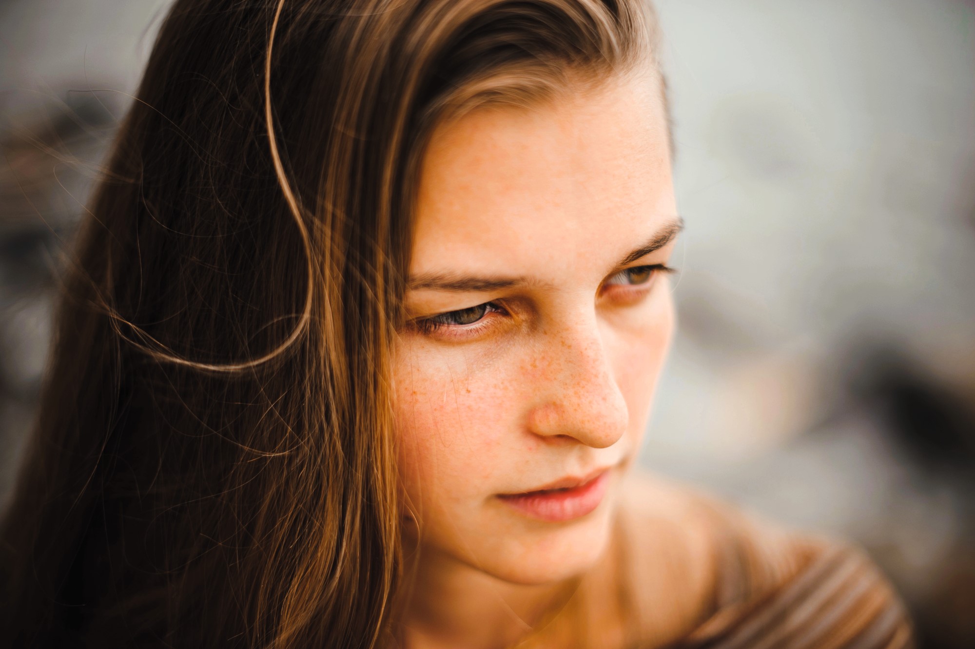 Close-up of a person with long brown hair, looking down and to the side. The background is blurred, highlighting their thoughtful expression and natural skin with light freckles.