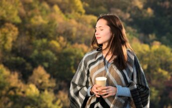 A woman with long brown hair stands outdoors wrapped in a plaid blanket, holding a yellow cup. Her eyes are closed, enjoying the sunlight. The background is a blurred view of trees with autumn foliage.