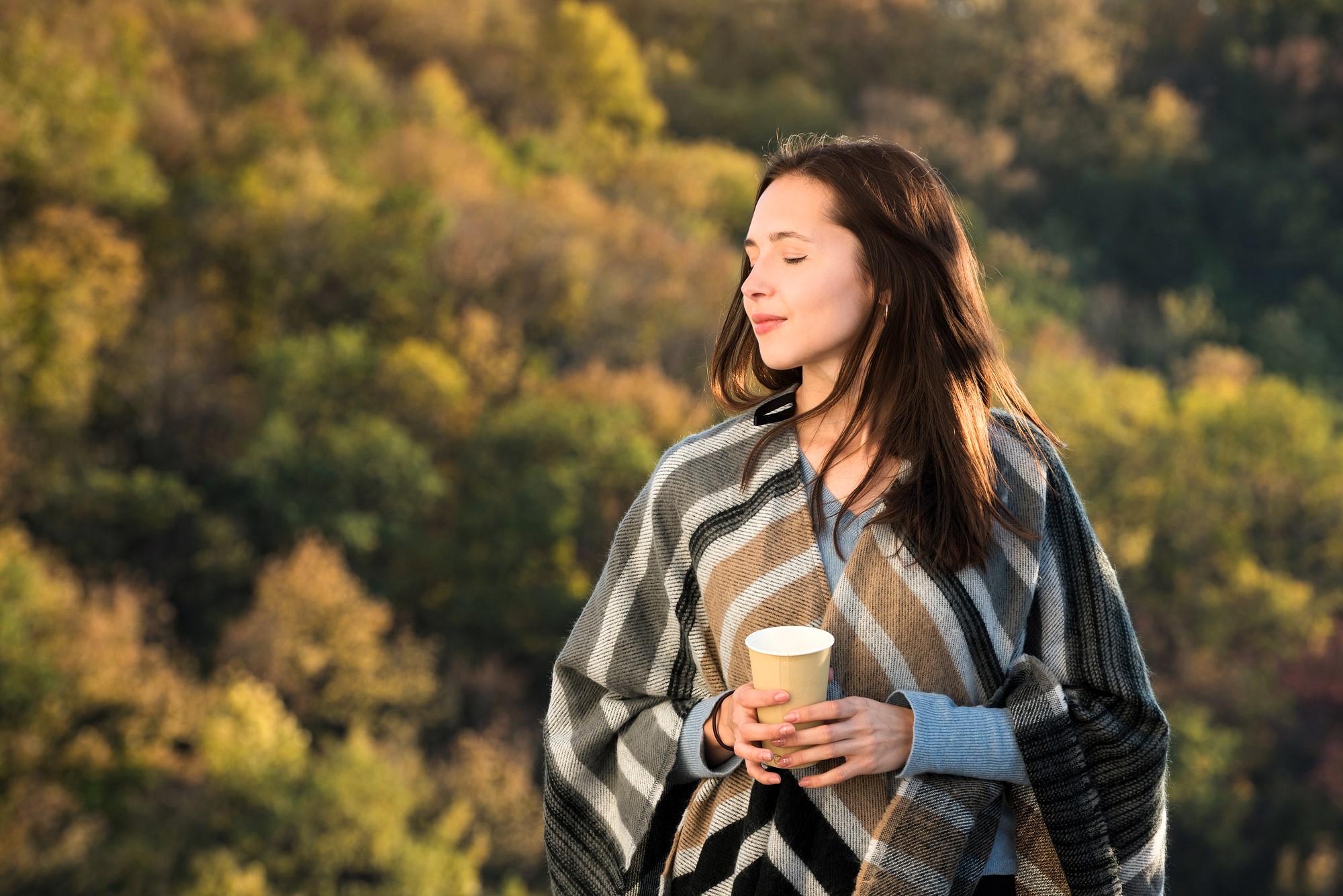 A woman with long brown hair stands outdoors wrapped in a plaid blanket, holding a yellow cup. Her eyes are closed, enjoying the sunlight. The background is a blurred view of trees with autumn foliage.