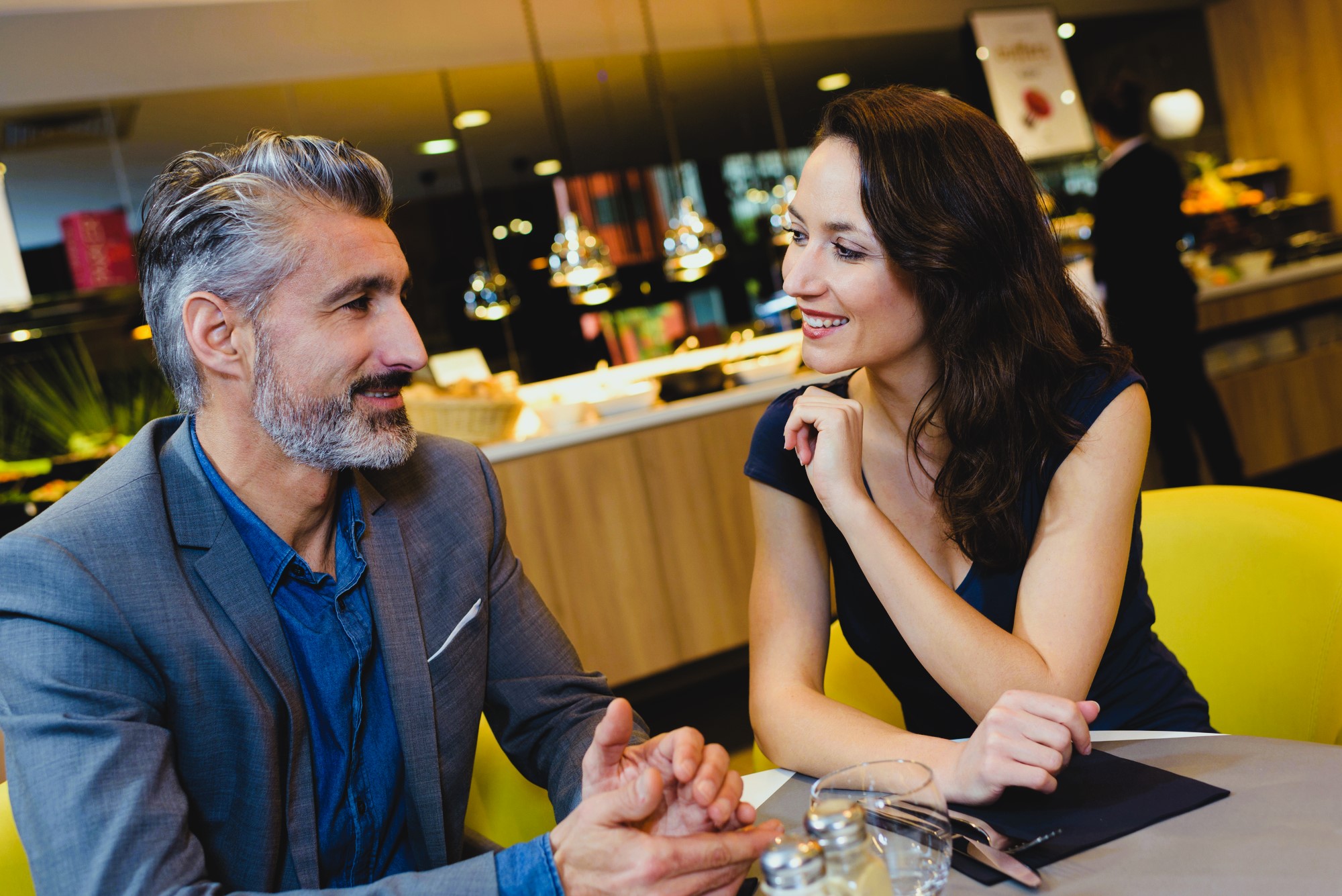 A man and woman are sitting at a table in a modern restaurant, engaged in conversation. The man has gray hair and a beard, wearing a gray suit. The woman has long brown hair and is wearing a black dress. The setting is warmly lit.