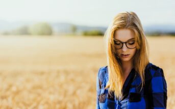 A woman with long blonde hair and glasses is standing outdoors in a field of wheat. She is wearing a blue and black checkered shirt and looking down. The background is a blurred landscape with a clear sky overhead.