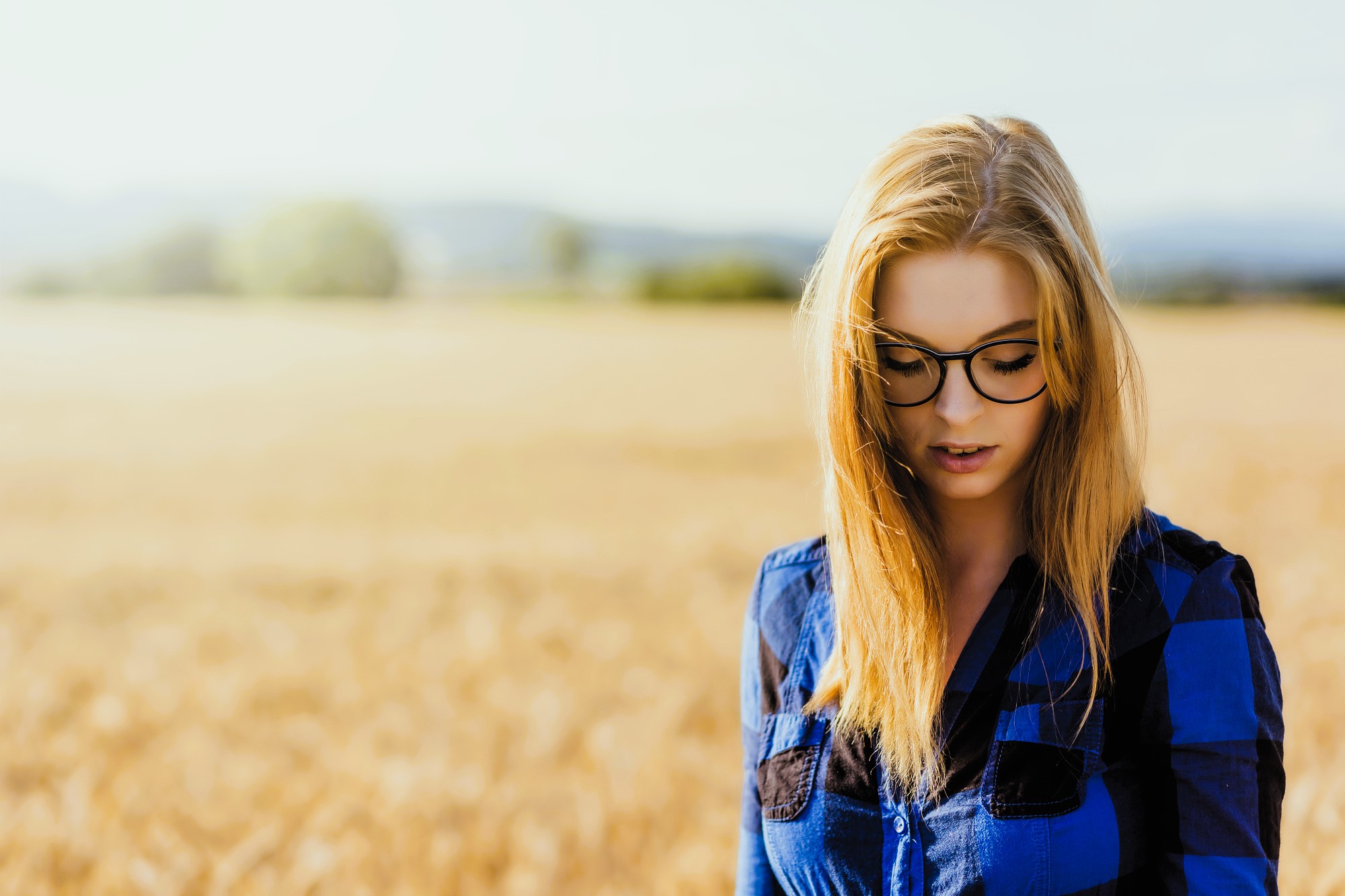 A woman with long blonde hair and glasses is standing outdoors in a field of wheat. She is wearing a blue and black checkered shirt and looking down. The background is a blurred landscape with a clear sky overhead.