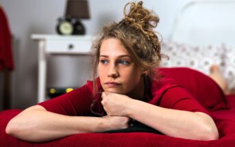 Woman with curly hair lying on a bed wearing a red shirt, resting her chin on her hands. She appears thoughtful, looking to the side. The room has a white bedside table with a lamp and alarm clock.