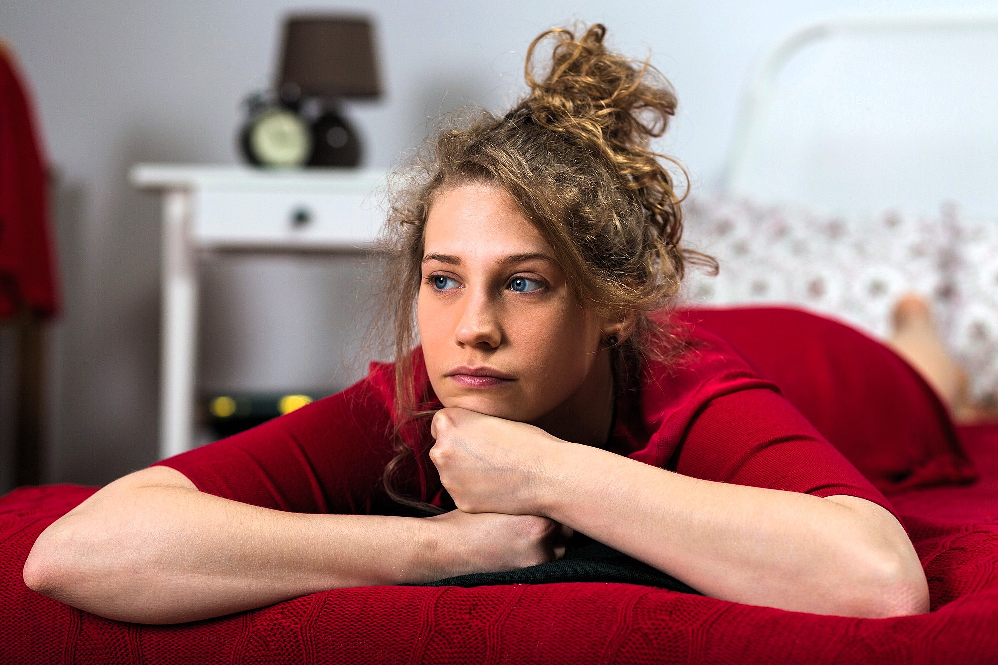 Woman with curly hair lying on a bed wearing a red shirt, resting her chin on her hands. She appears thoughtful, looking to the side. The room has a white bedside table with a lamp and alarm clock.