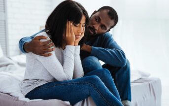 A man sitting on a bed comforts a woman who looks upset, with her head resting on her hands. They are both casually dressed in jeans and long-sleeve tops. The room is softly lit with a bright, blurred background.