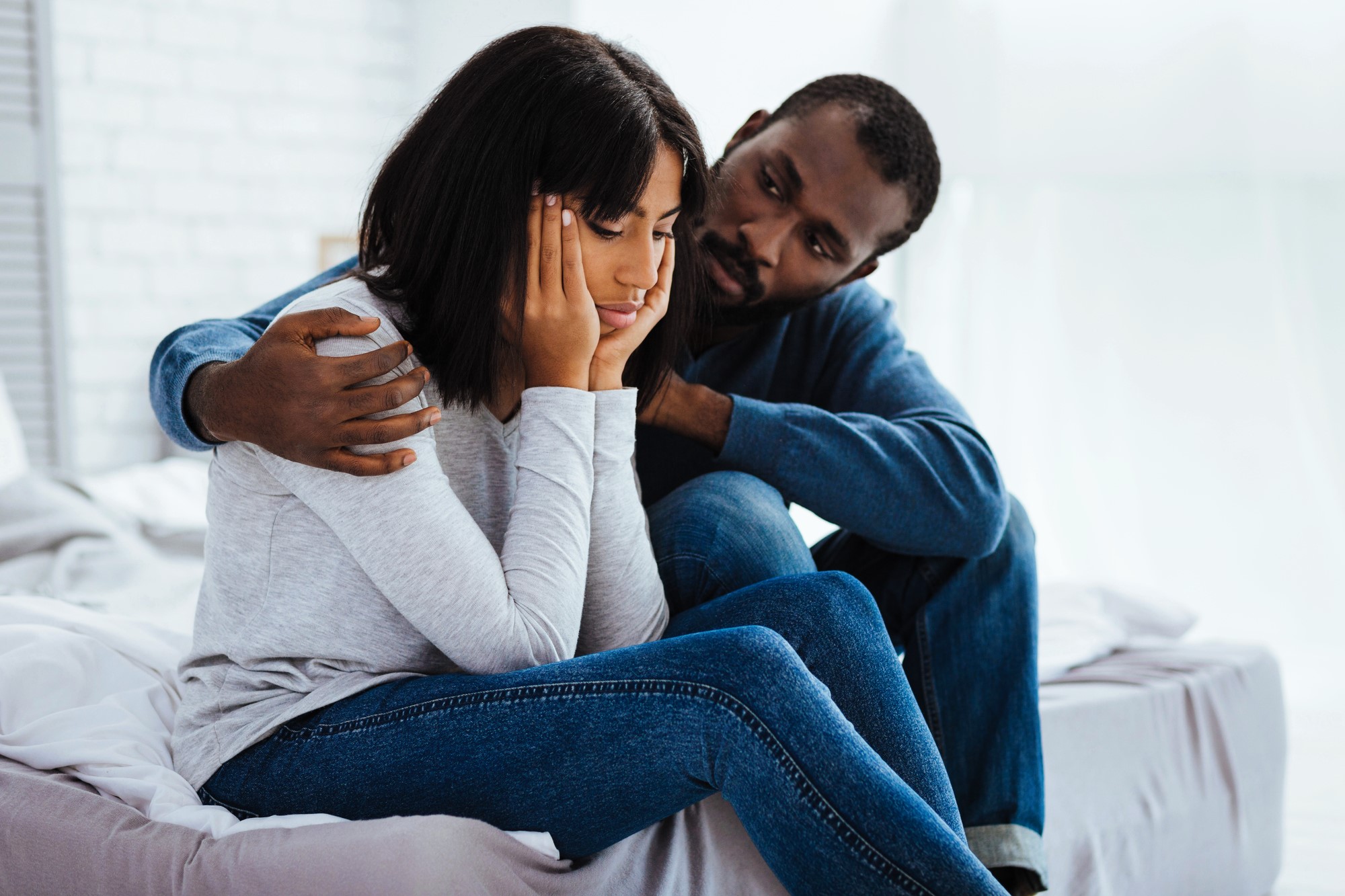 A man sitting on a bed comforts a woman who looks upset, with her head resting on her hands. They are both casually dressed in jeans and long-sleeve tops. The room is softly lit with a bright, blurred background.