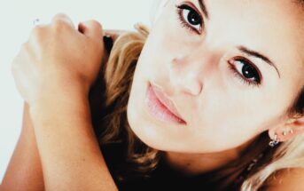 A close-up of a person with light hair and earrings looking directly at the camera. The background is white, and their arm is resting near their face. The image has a high-contrast and slightly warm tone.