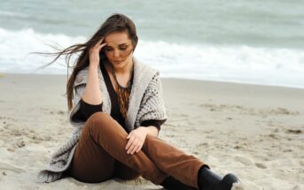 A woman with long hair sits on a sandy beach, wearing brown pants, a knitted sweater, and black boots. She has her eyes closed and holds her hand to her forehead. The ocean is visible in the background under an overcast sky.