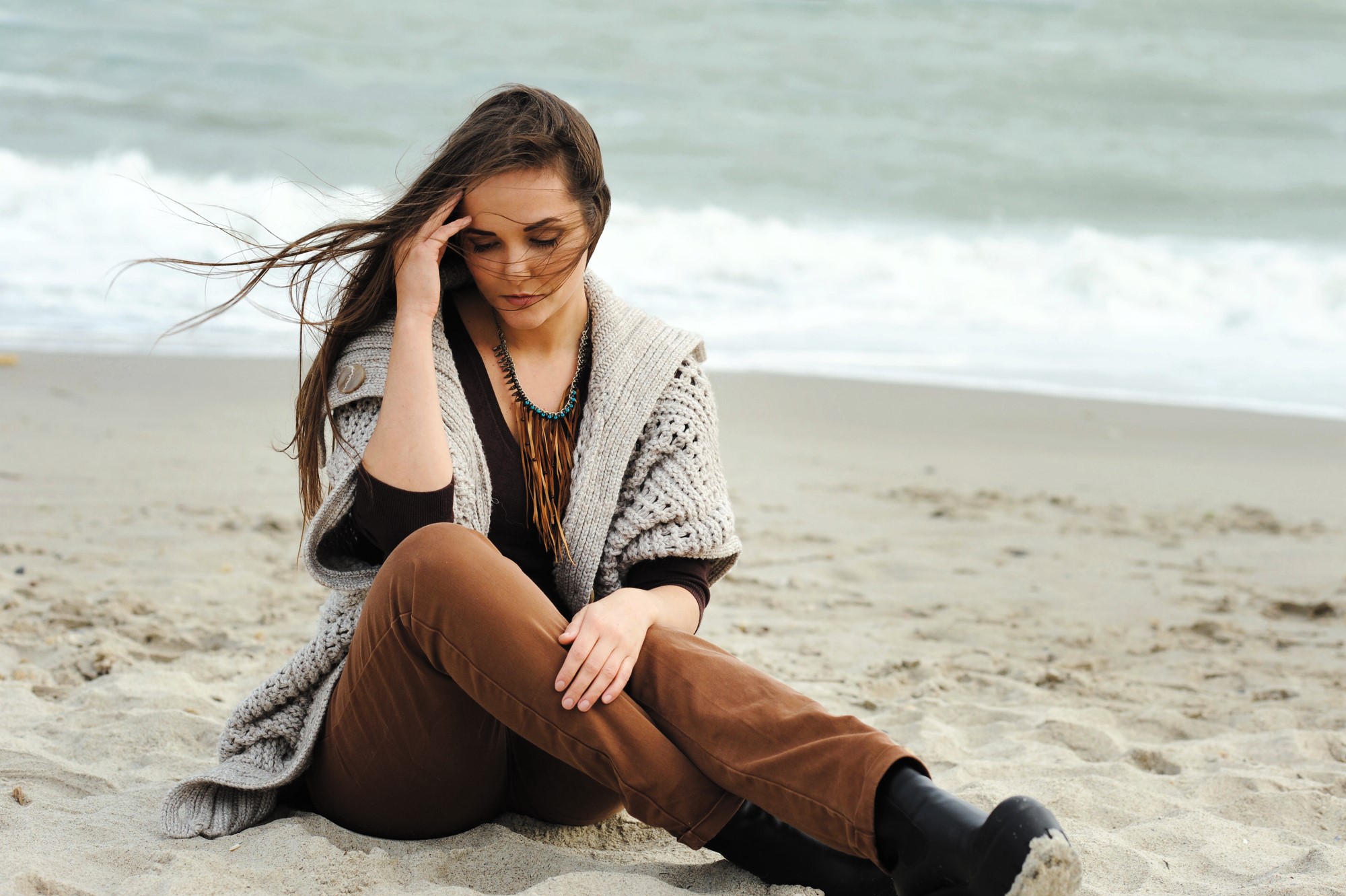 A woman with long hair sits on a sandy beach, wearing brown pants, a knitted sweater, and black boots. She has her eyes closed and holds her hand to her forehead. The ocean is visible in the background under an overcast sky.