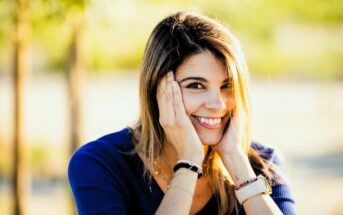 A woman with long hair smiles warmly, resting her hands on her cheeks. She is wearing a blue top and has a watch on her wrist. The background is sunny and blurred, suggesting an outdoor setting.