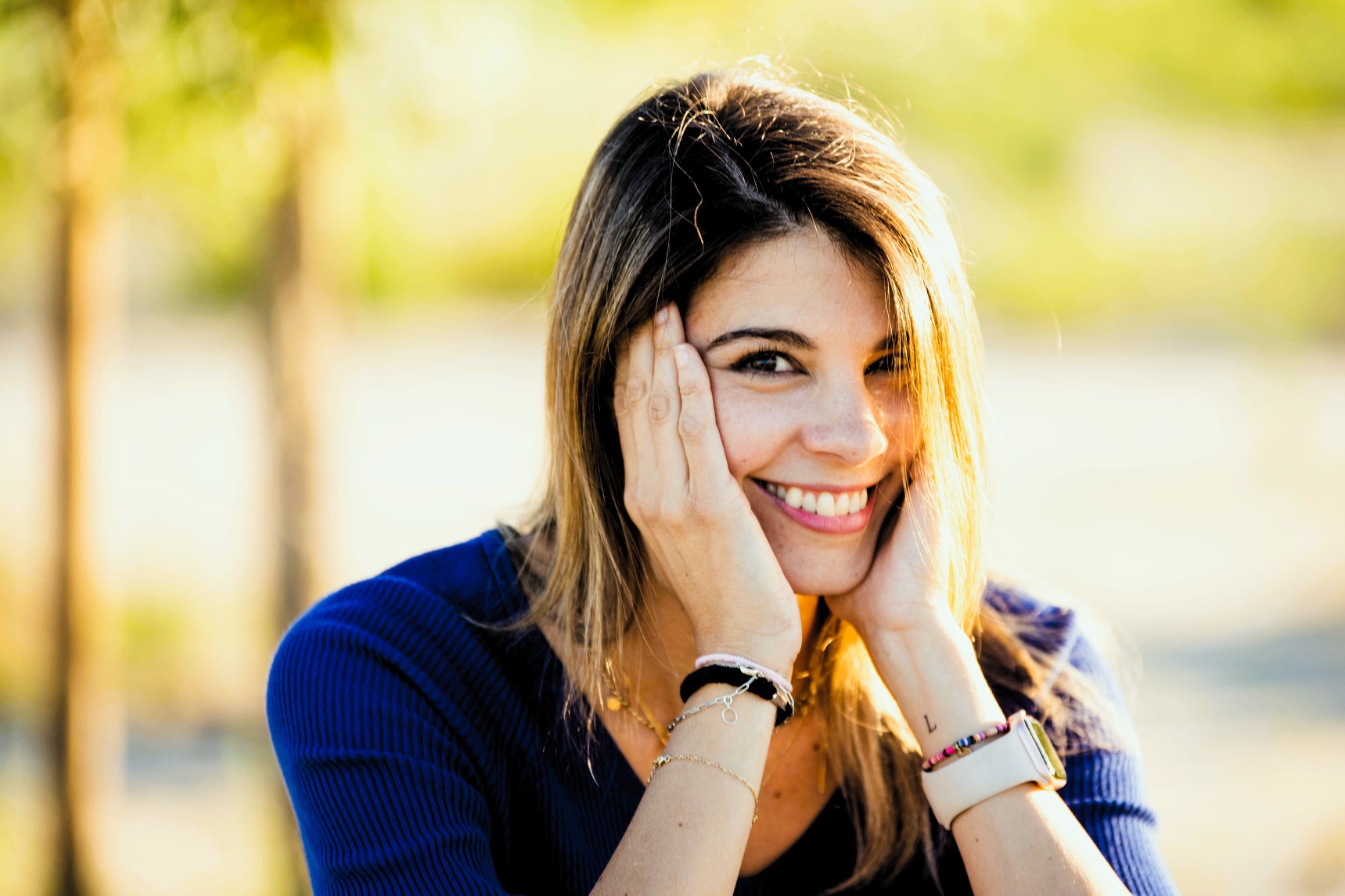 A woman with long hair smiles warmly, resting her hands on her cheeks. She is wearing a blue top and has a watch on her wrist. The background is sunny and blurred, suggesting an outdoor setting.