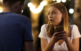 Woman with long hair holding a phone looks intently at a person across from her. She is wearing a white shirt. The background is softly lit with blurred lights, creating a warm ambiance.