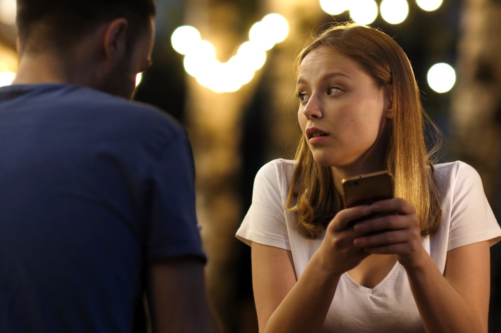 Woman with long hair holding a phone looks intently at a person across from her. She is wearing a white shirt. The background is softly lit with blurred lights, creating a warm ambiance.