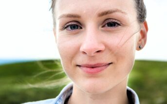 A person with light skin and blue eyes, smiling softly, is shown in close-up. They stand outdoors with grassy hills in the background. Their hair is tied back and they wear stud earrings. The lighting is bright and natural.