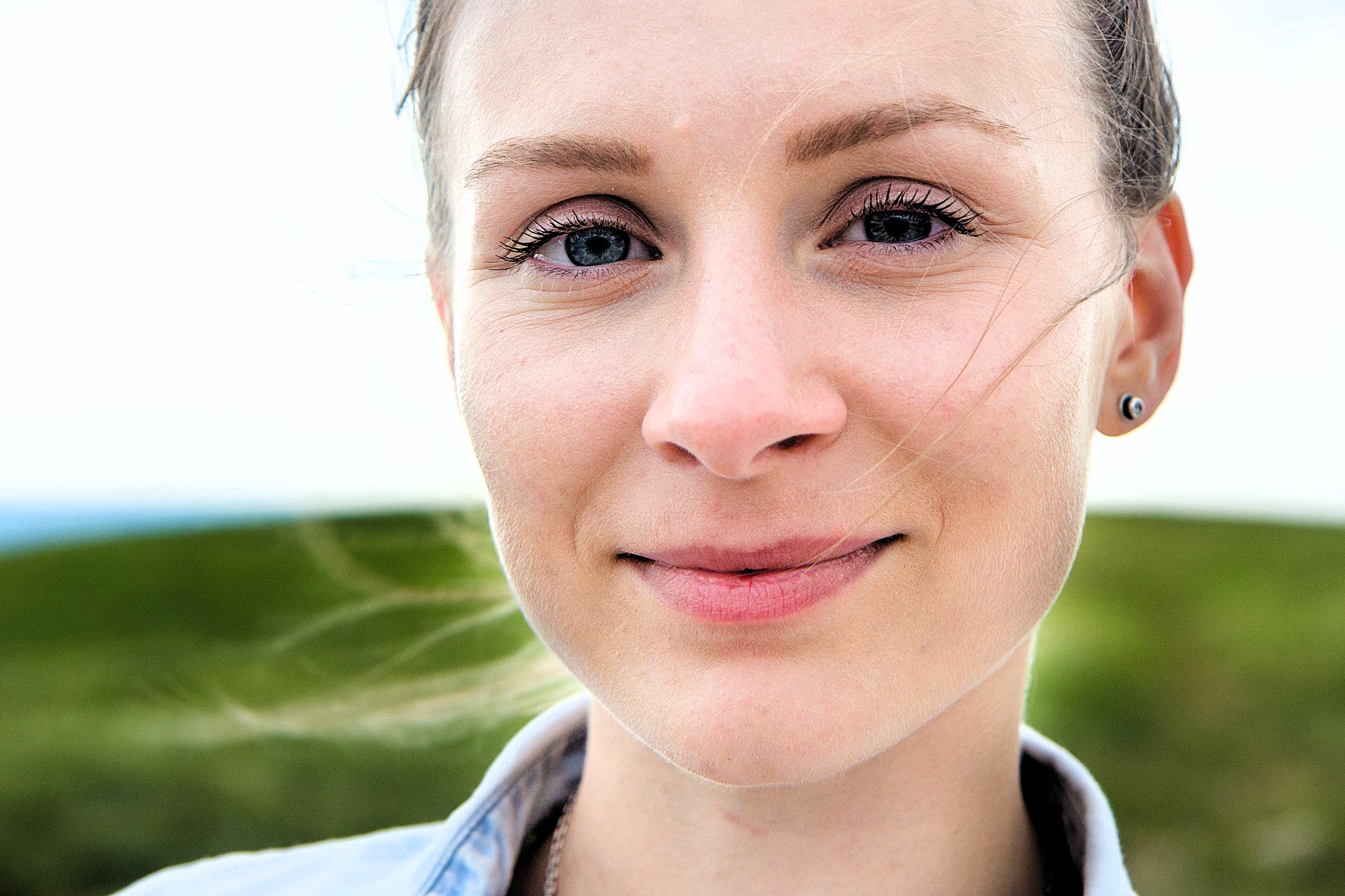 A person with light skin and blue eyes, smiling softly, is shown in close-up. They stand outdoors with grassy hills in the background. Their hair is tied back and they wear stud earrings. The lighting is bright and natural.