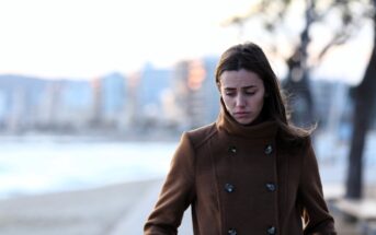 A woman in a brown coat stands by a beach, looking downwards with a thoughtful expression. Blurred city buildings and trees are in the background, with soft, natural lighting.