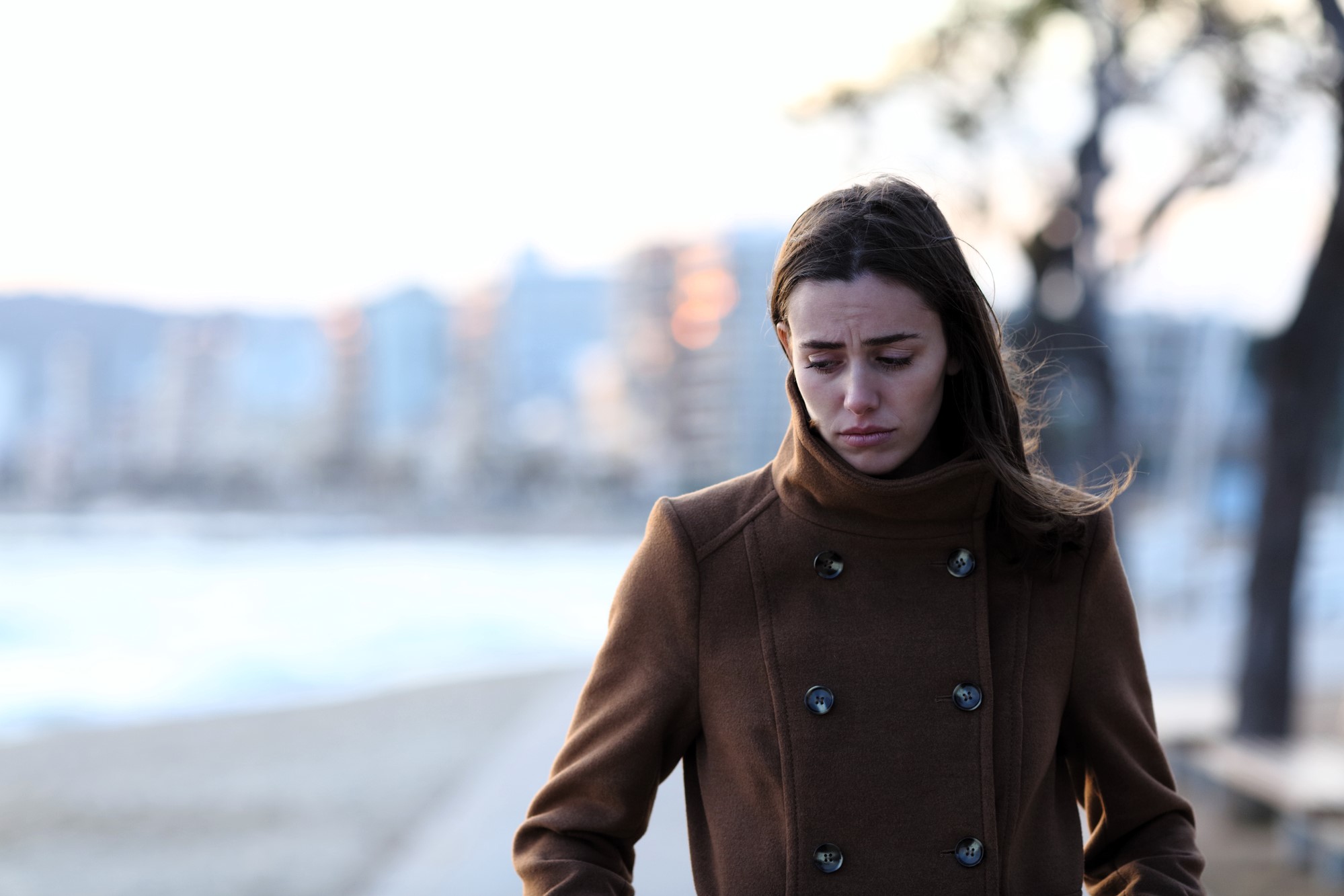 A woman in a brown coat stands by a beach, looking downwards with a thoughtful expression. Blurred city buildings and trees are in the background, with soft, natural lighting.