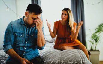 A man in a denim shirt sits on a bed, resting his forehead in his hand, looking distressed. A woman in an orange dress sits beside him with arms raised, expressing concern. The room is softly lit, with a plant and curtains in the background.