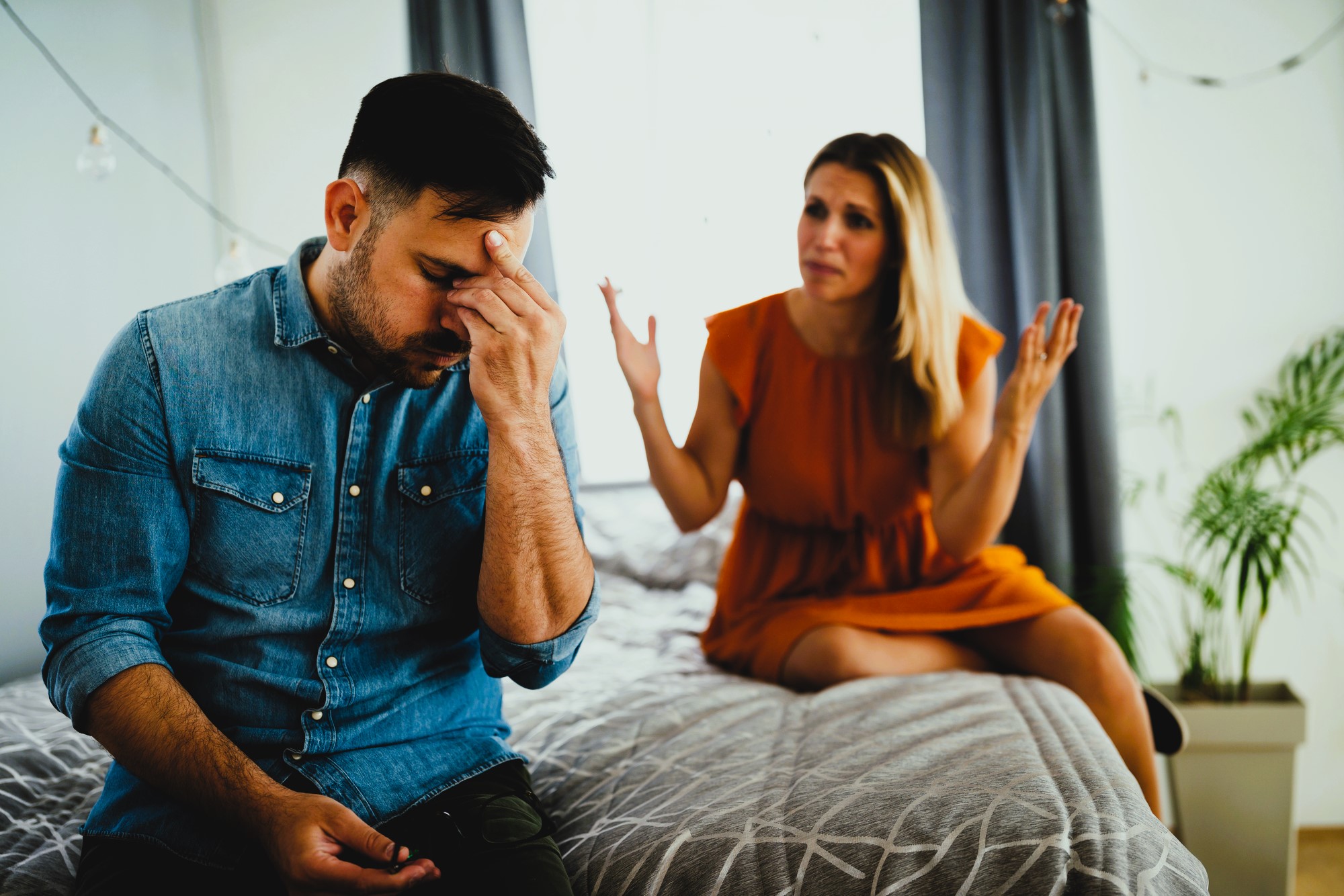 A man in a denim shirt sits on a bed, resting his forehead in his hand, looking distressed. A woman in an orange dress sits beside him with arms raised, expressing concern. The room is softly lit, with a plant and curtains in the background.