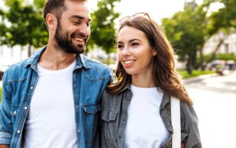 A man and woman walking outdoors on a sunny day. Both are smiling and casually dressed, with the man in a denim shirt and the woman in a gray shirt with sunglasses on her head. Green trees and a street are visible in the background.