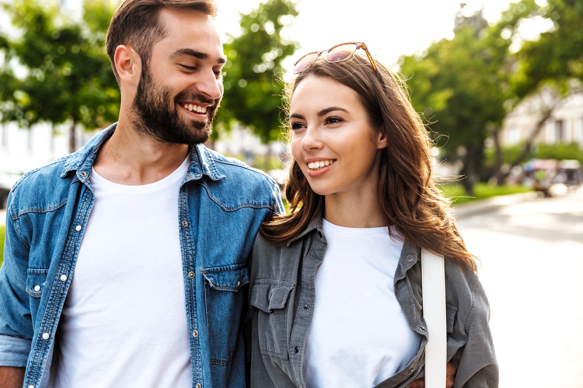 A man and woman walking outdoors on a sunny day. Both are smiling and casually dressed, with the man in a denim shirt and the woman in a gray shirt with sunglasses on her head. Green trees and a street are visible in the background.