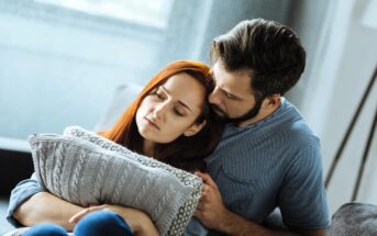 A couple sits close on a couch. The woman, with long red hair, looks pensive while holding a knitted pillow. The man, with short dark hair and a beard, tenderly leans against her, offering comfort. They both wear casual clothing.