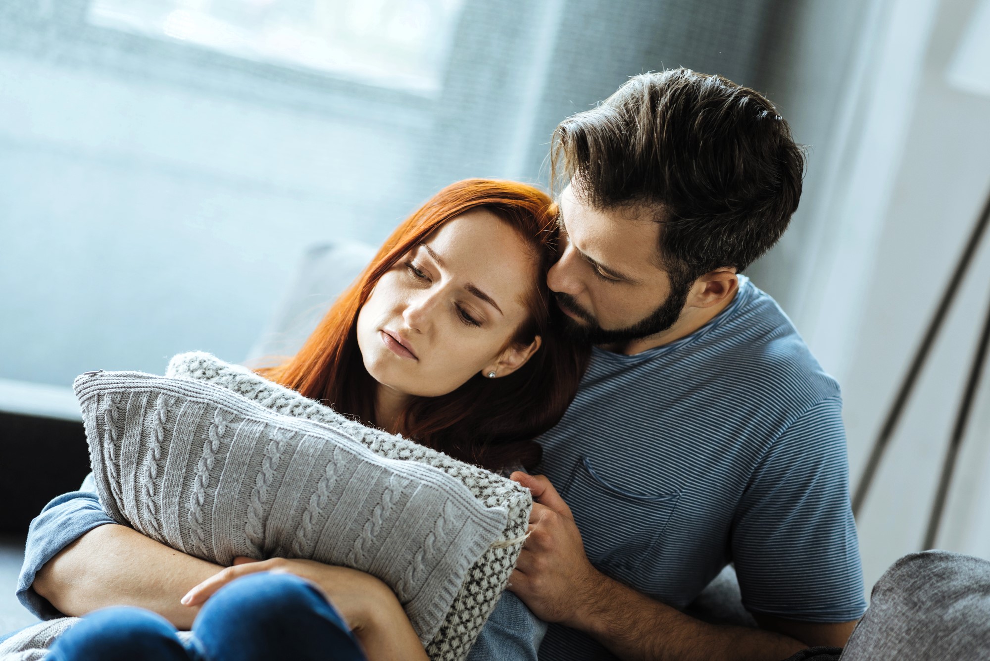 A couple sits close on a couch. The woman, with long red hair, looks pensive while holding a knitted pillow. The man, with short dark hair and a beard, tenderly leans against her, offering comfort. They both wear casual clothing.