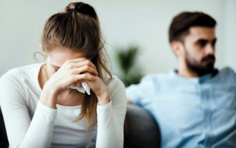 A woman in a white shirt, appearing upset, sits with her head in her hands holding a tissue. A man in a blue shirt sits beside her, looking away with a stern expression. Both are on a sofa in a softly lit room.