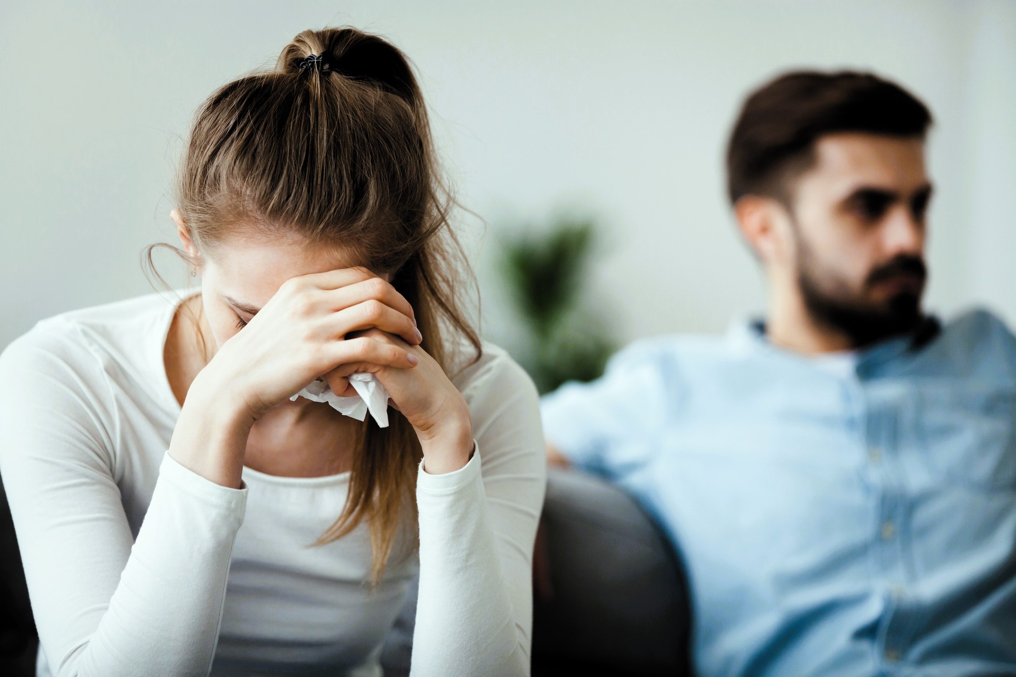 A woman in a white shirt, appearing upset, sits with her head in her hands holding a tissue. A man in a blue shirt sits beside her, looking away with a stern expression. Both are on a sofa in a softly lit room.