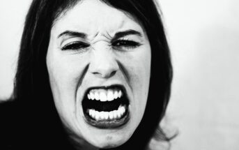 Close-up black and white image of a person displaying an intense expression, with their mouth open wide showing teeth. Their eyes are slightly squinted, and they appear to be shouting or expressing strong emotion.