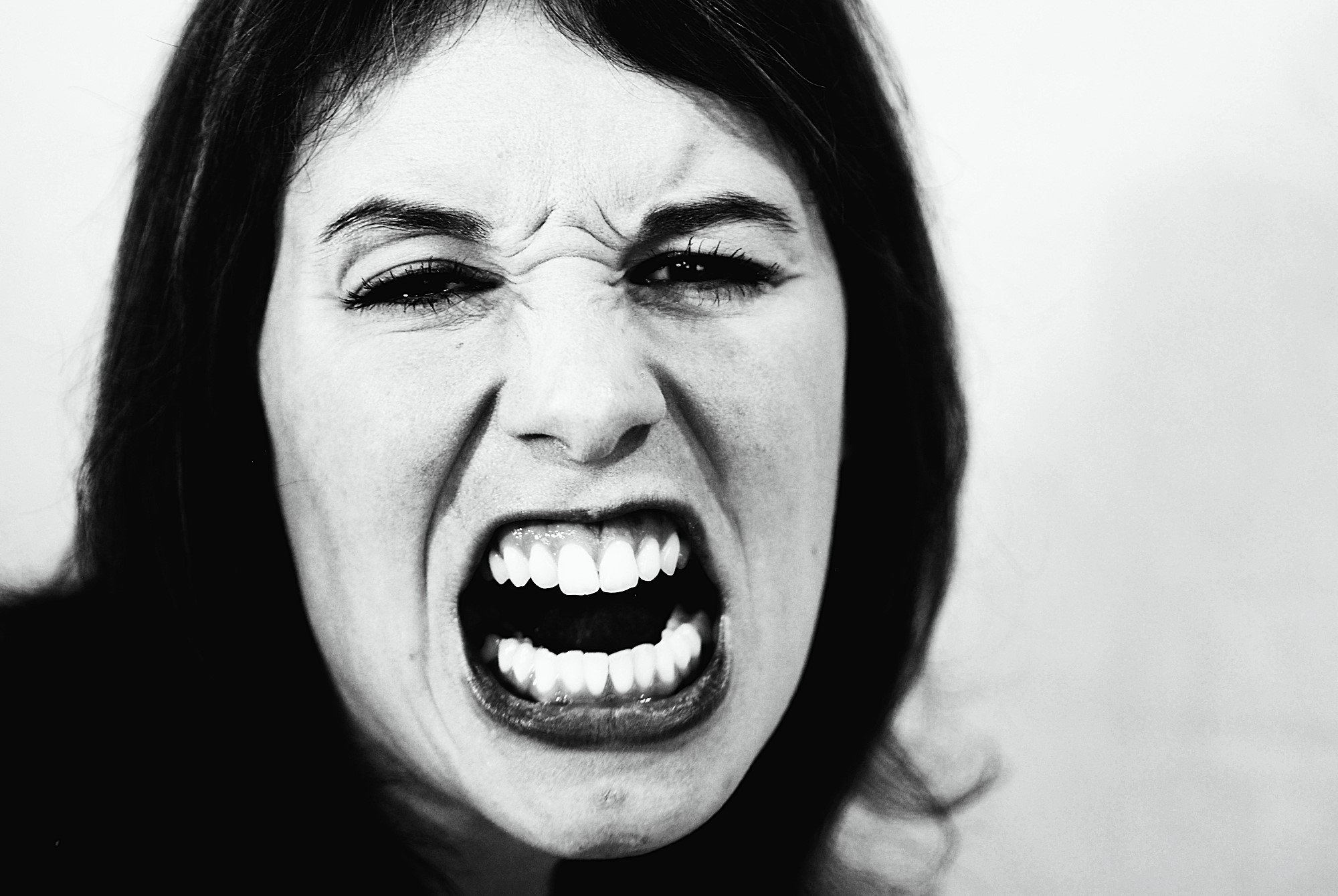 Close-up black and white image of a person displaying an intense expression, with their mouth open wide showing teeth. Their eyes are slightly squinted, and they appear to be shouting or expressing strong emotion.