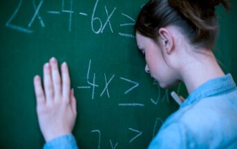 A person with brown hair, wearing a denim shirt, rests their forehead against a green chalkboard filled with algebraic equations. They appear frustrated or deep in thought.