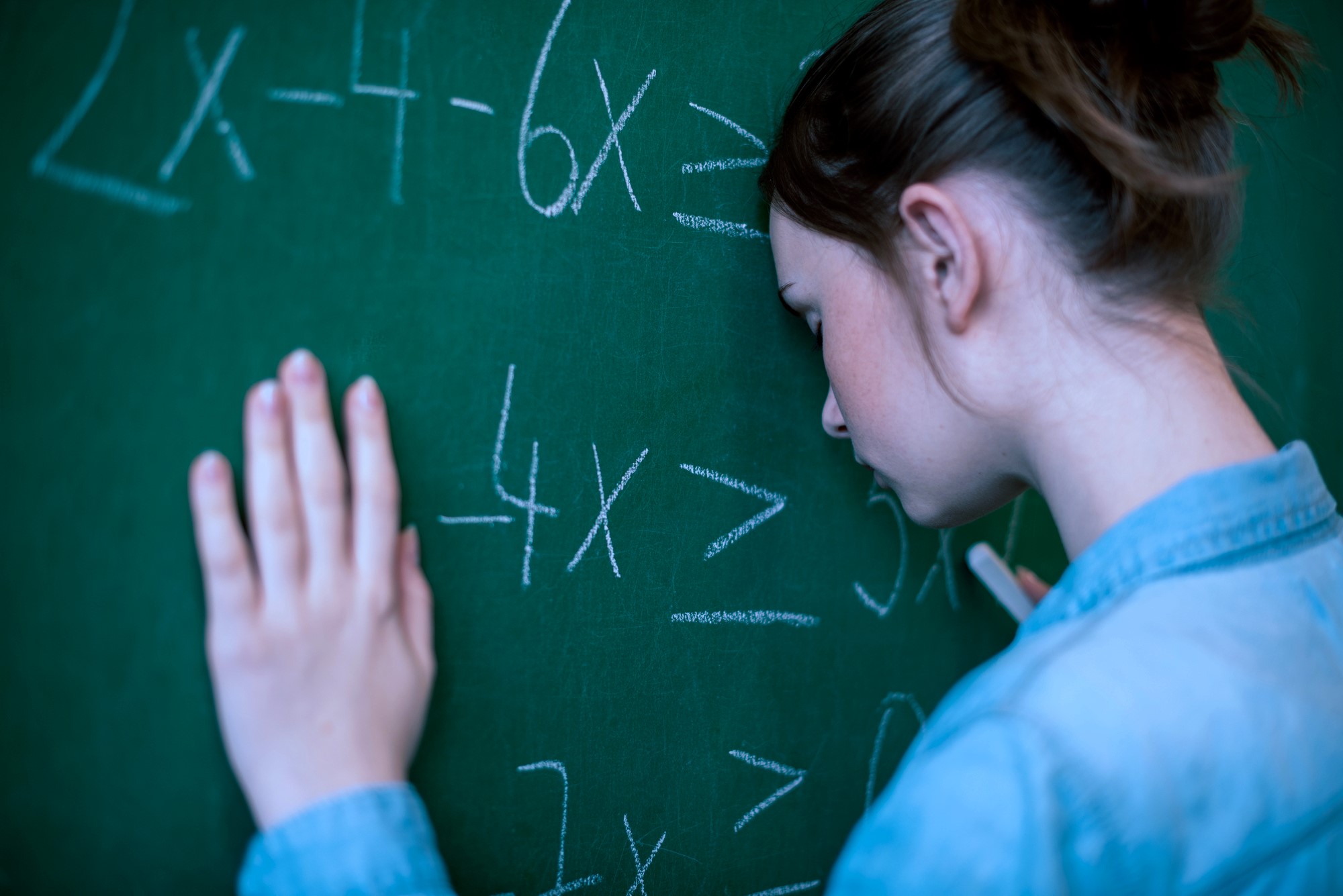 A person with brown hair, wearing a denim shirt, rests their forehead against a green chalkboard filled with algebraic equations. They appear frustrated or deep in thought.