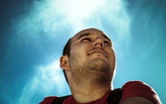 A man in a red shirt looks up at the sky, smiling slightly. The photo is taken from a low angle, with a clear, bright blue sky in the background. The sunlight creates a soft halo effect around his head.