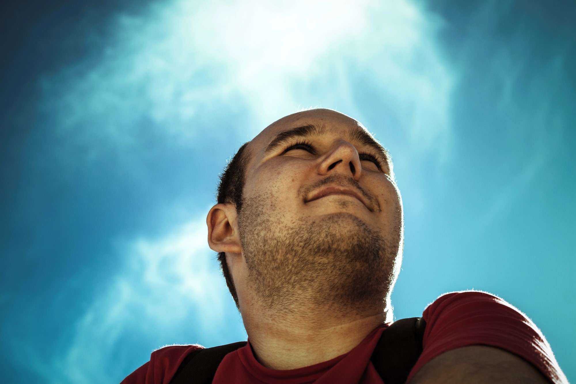 A man in a red shirt looks up at the sky, smiling slightly. The photo is taken from a low angle, with a clear, bright blue sky in the background. The sunlight creates a soft halo effect around his head.