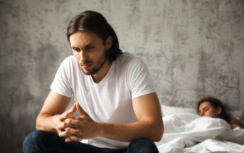 A man with long hair and a beard sits on the edge of a bed, deep in thought, wearing a white t-shirt and jeans. Behind him, a person is sleeping under white sheets. The room has a textured gray wall.