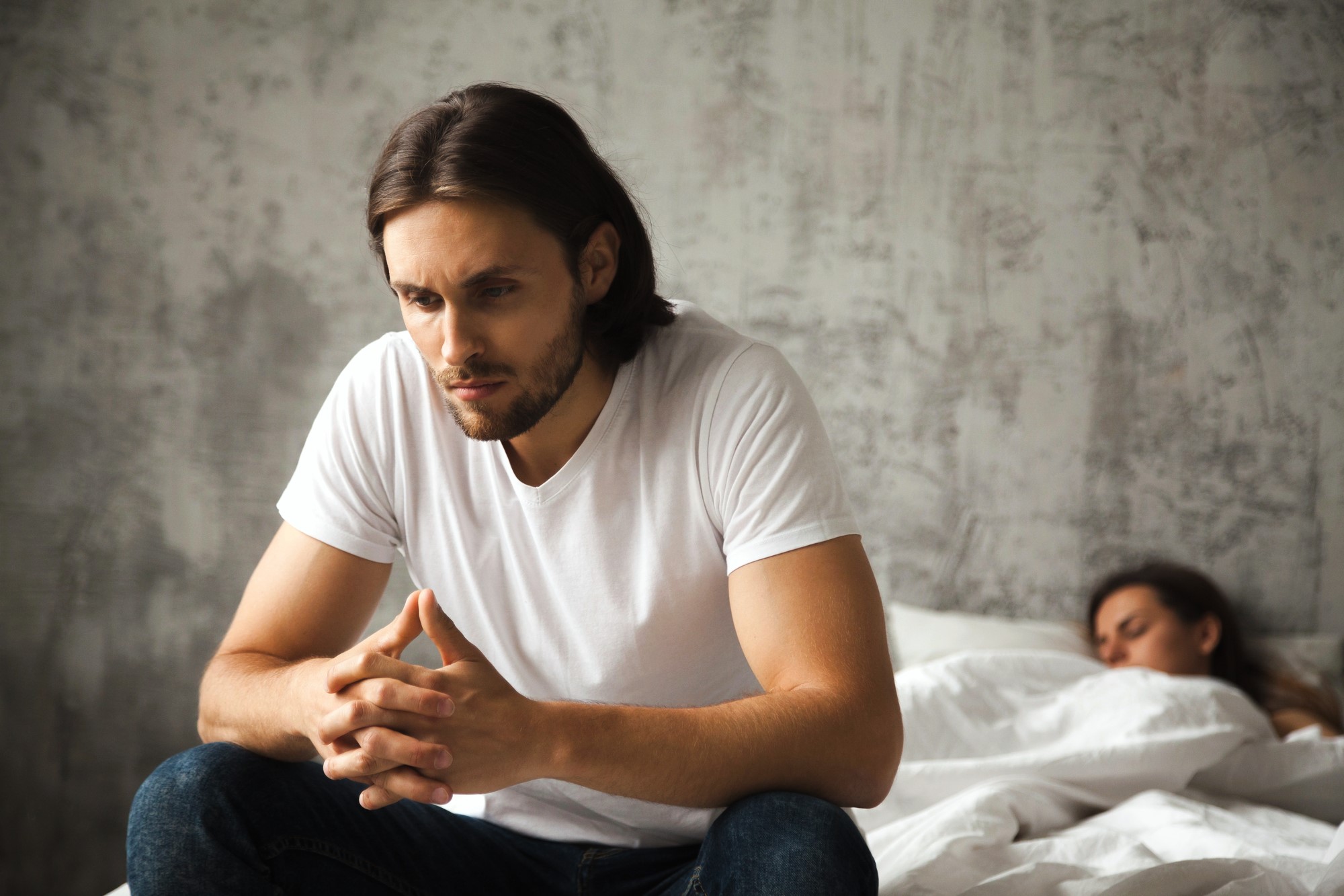 A man with long hair and a beard sits on the edge of a bed, deep in thought, wearing a white t-shirt and jeans. Behind him, a person is sleeping under white sheets. The room has a textured gray wall.