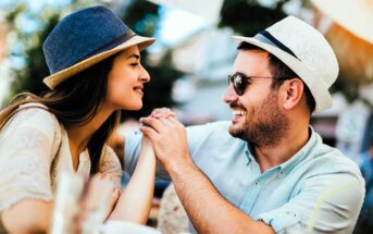 A smiling couple sits at an outdoor café. The woman with long hair wears a blue hat, and the man with a beard wears sunglasses and a white hat. They hold hands and face each other, surrounded by blurred cups and greenery in the background.