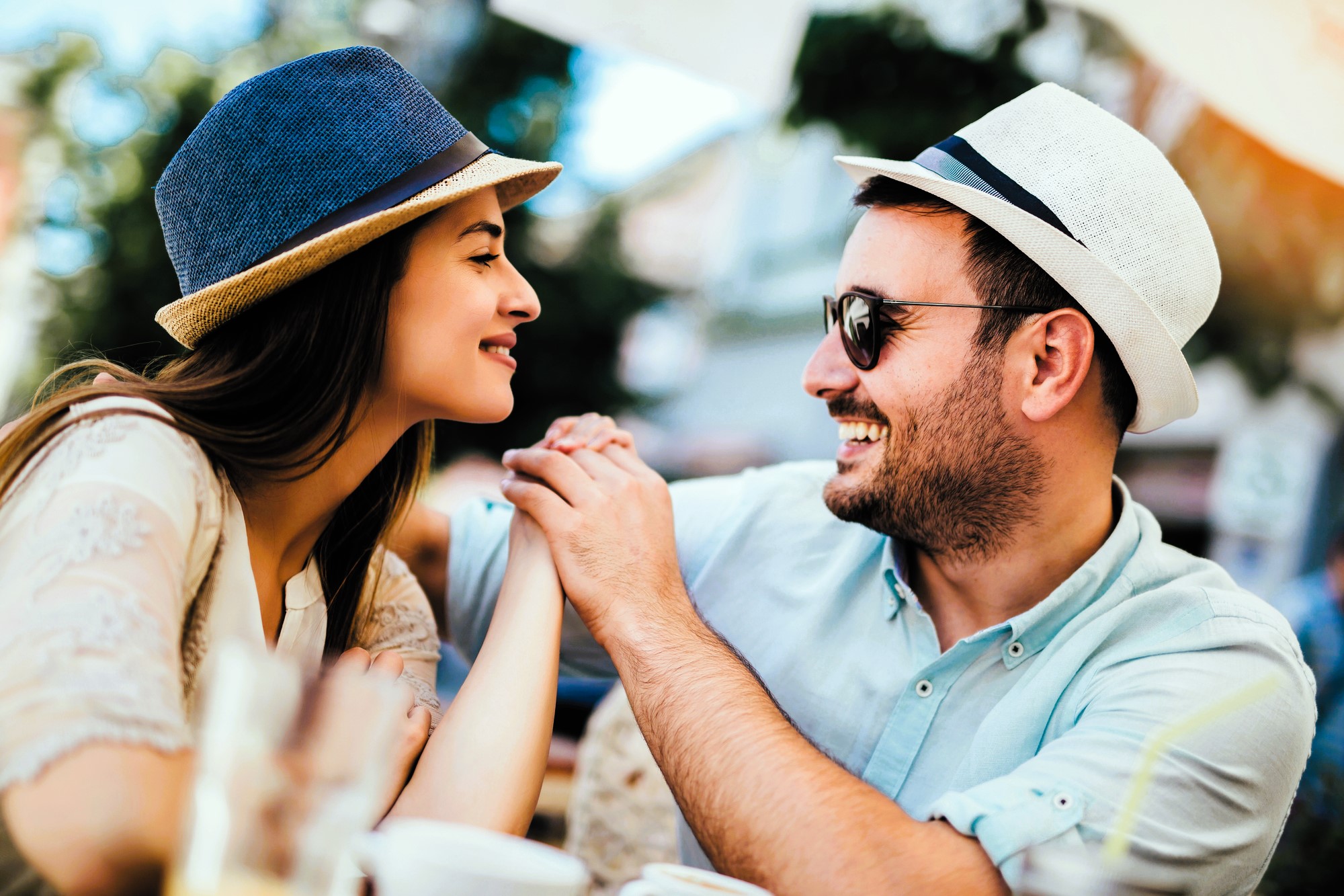 A smiling couple sits at an outdoor café. The woman with long hair wears a blue hat, and the man with a beard wears sunglasses and a white hat. They hold hands and face each other, surrounded by blurred cups and greenery in the background.