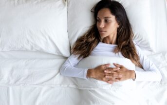 A woman with long dark hair lies in bed on her back, looking to the side. She is under a white blanket, wearing a white top, with her hands clasped on her abdomen. The setting is bright and minimalistic.