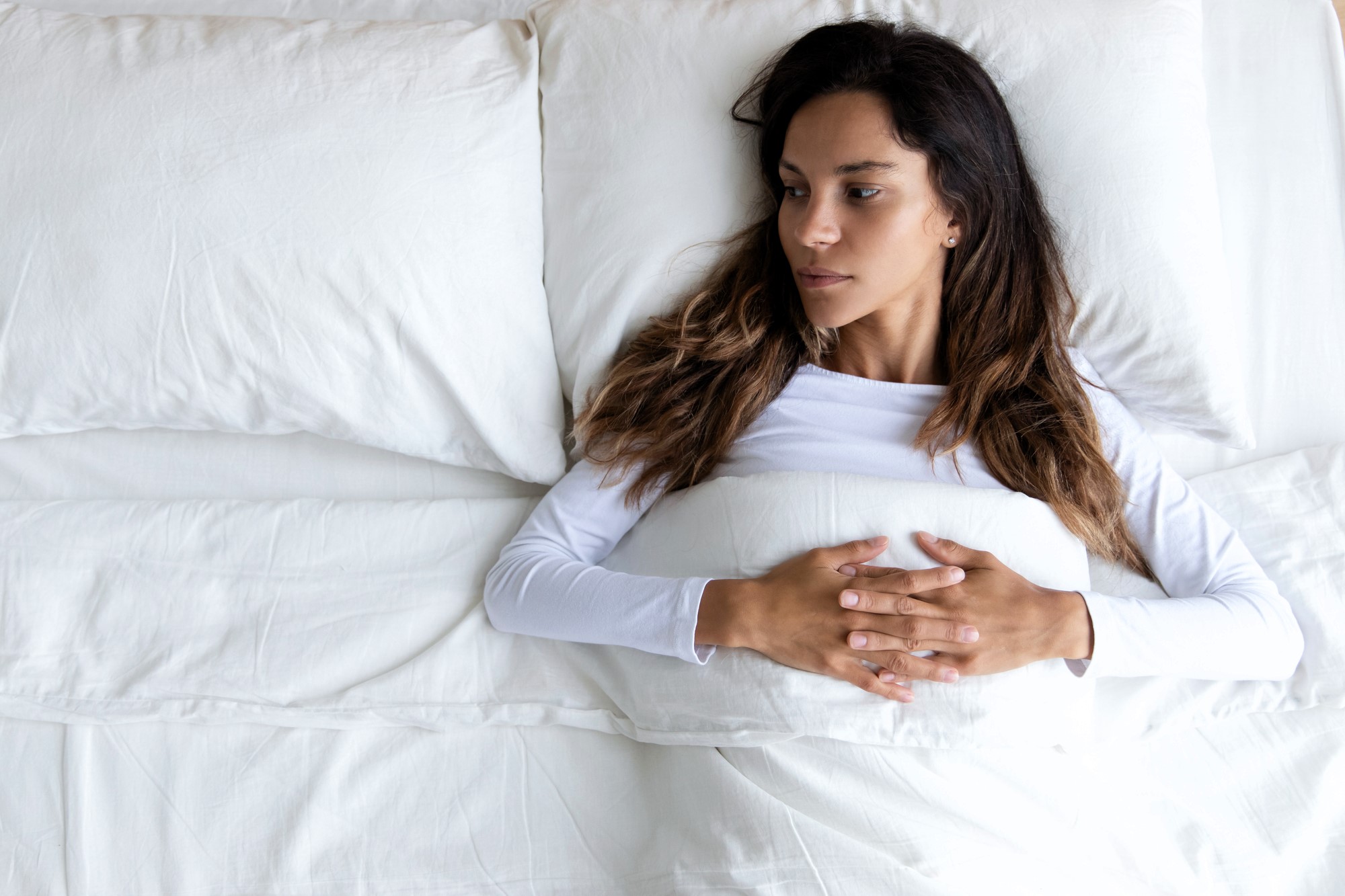 A woman with long dark hair lies in bed on her back, looking to the side. She is under a white blanket, wearing a white top, with her hands clasped on her abdomen. The setting is bright and minimalistic.