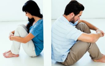 A man and woman sit back-to-back on the floor, separated by a wall. Both have their knees drawn up, arms wrapped around them, looking down. The woman is wearing a blue top and white pants; the man is in a light blue shirt and beige pants.