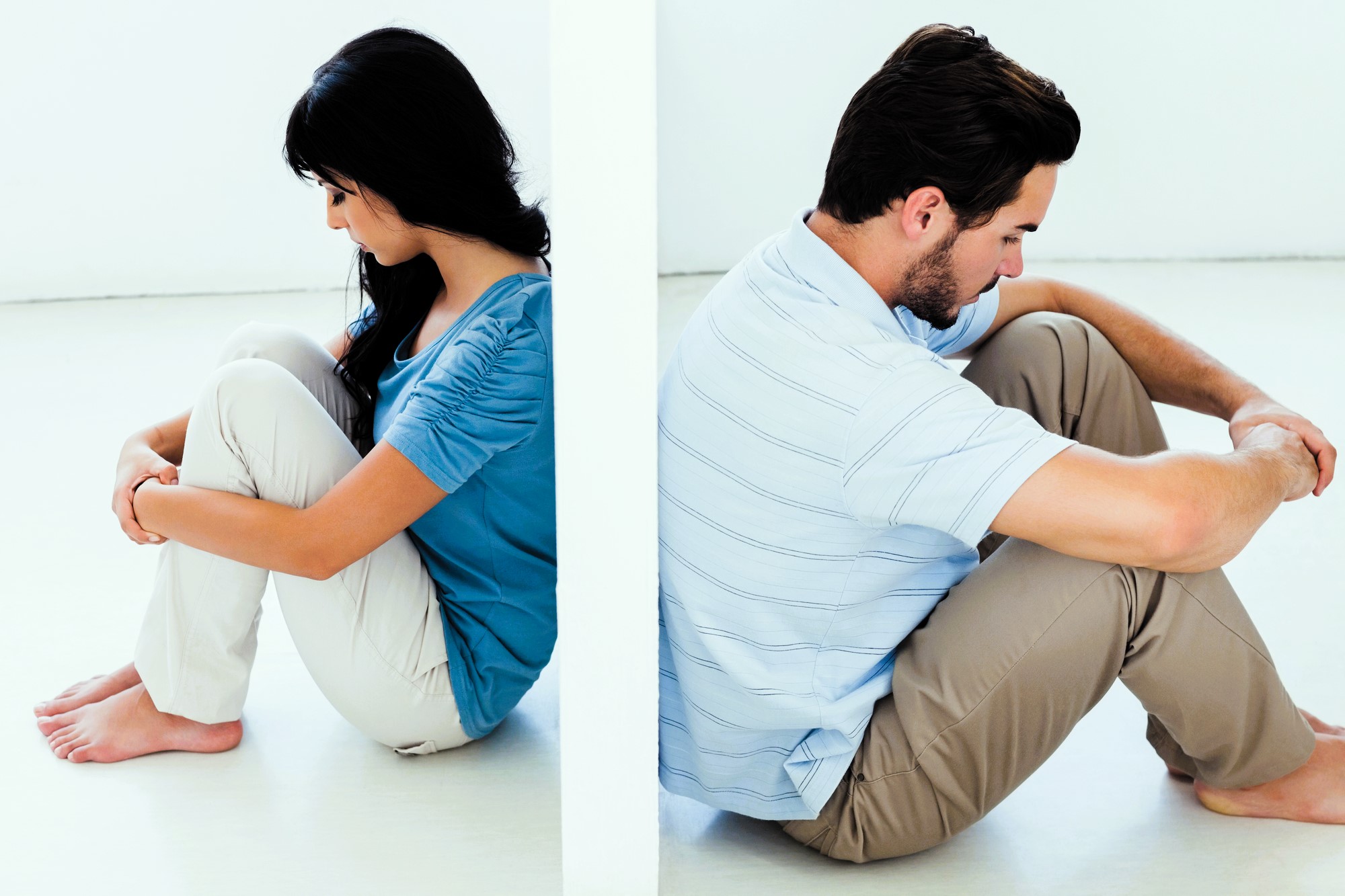 A man and woman sit back-to-back on the floor, separated by a wall. Both have their knees drawn up, arms wrapped around them, looking down. The woman is wearing a blue top and white pants; the man is in a light blue shirt and beige pants.