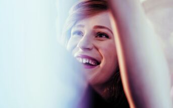 A person with light brown hair is smiling widely and looking upward. The image has a soft light effect, giving a warm and cheerful ambiance. Arms are raised, partially visible in the foreground.