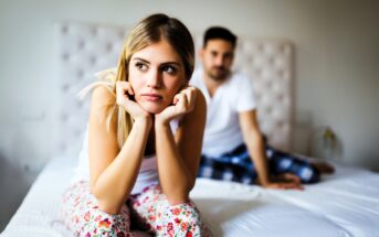 A woman with long blonde hair in floral pajama pants sits on a bed, resting her chin on her hands with a thoughtful expression. A man in a white shirt and plaid pants sits behind her, looking in her direction.