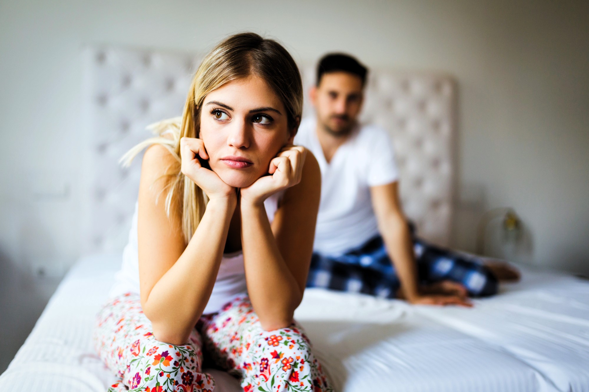 A woman with long blonde hair in floral pajama pants sits on a bed, resting her chin on her hands with a thoughtful expression. A man in a white shirt and plaid pants sits behind her, looking in her direction.