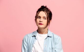 Woman with dark hair in a bun, wearing a light denim shirt over a white t-shirt, standing against a pink background, looking to the side with a neutral expression.